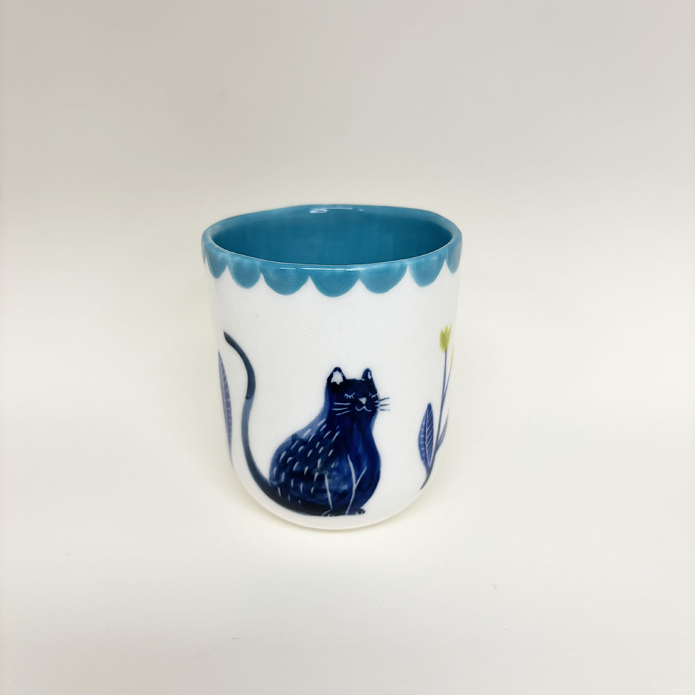 tasse en porcelaine, l'intérieur est bleu ciel avec un liseré bleu ciel à l'extérieur. un chat bleu foncé est dessiné il y a également, des fleur au pétales jaunes sur les coté du chat