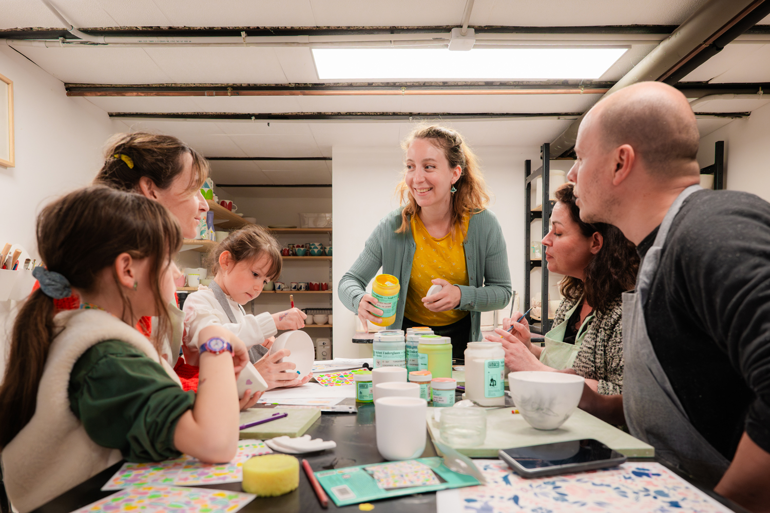 atelier peinture sur céramique. plusieurs personnes sont assis autour de la table enfant et adulte. Il me regarde je suis entrain de leurs expliqué l'utilisation de engobes. Je tiens u pot d'engobe jaune en main
